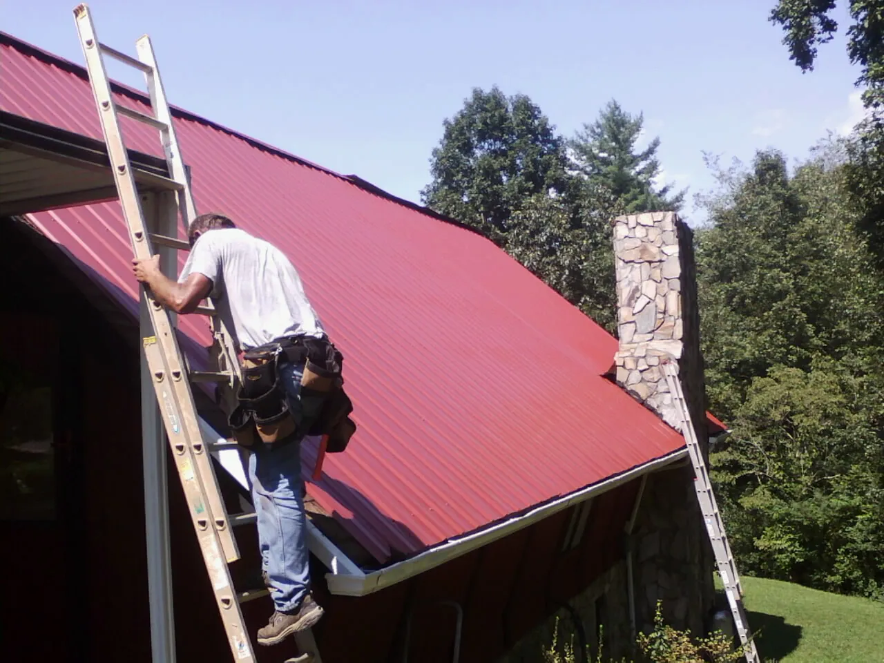 Roofer on a ladder beside a red metal roof and stone chimney — Soffit & Fascia Repair by Roofing Brothers in Fairplay